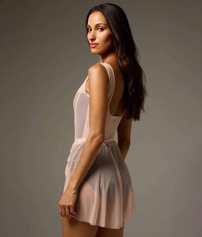 A woman modeling a pale blush pink sleeveless mini dress with a sheer mesh bodice and a flared skirt, photographed against a plain taupe-gray studio background.