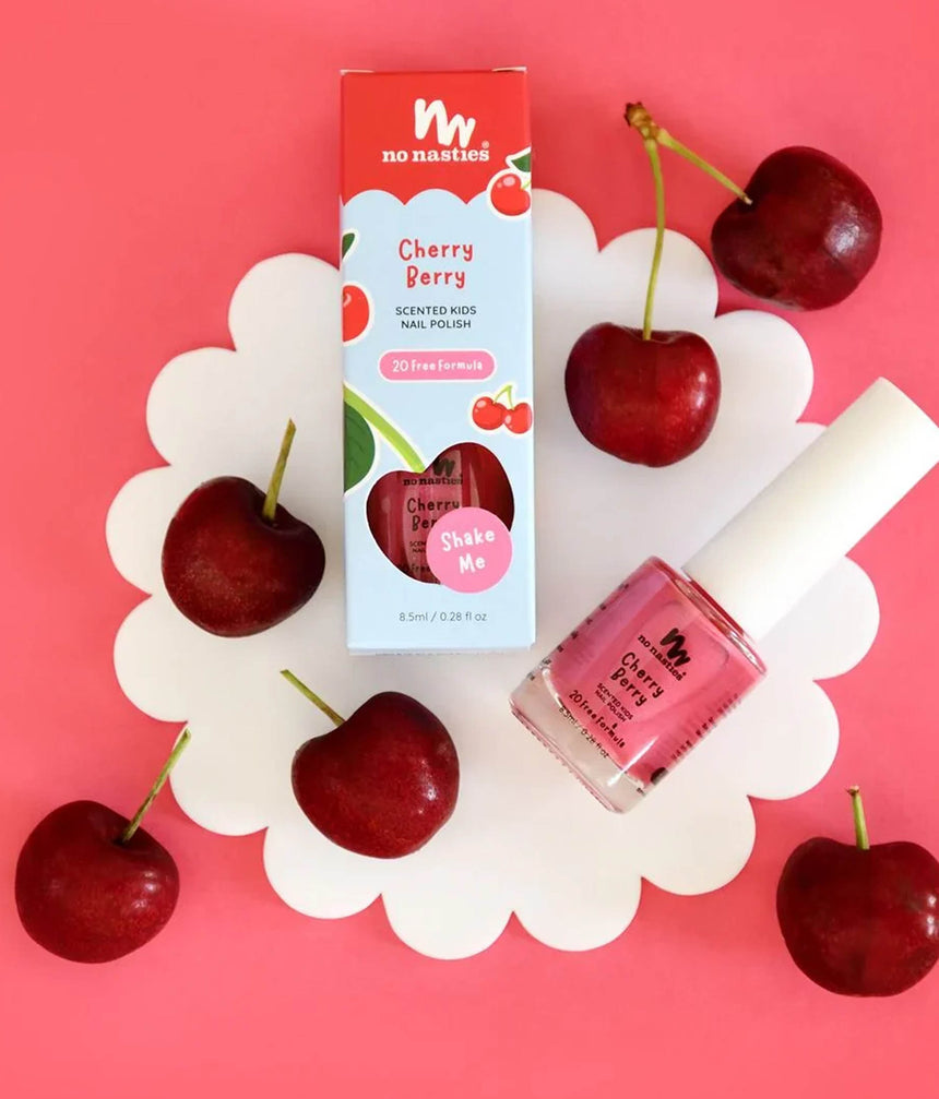 A bottle and box of No Nasties Cherry Berry Scented Kid Nail Polish arranged on a white scalloped tray with fresh cherries on a pink background.