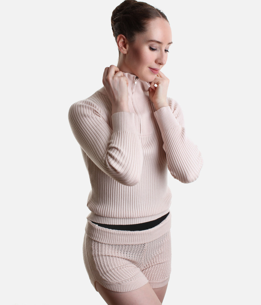 A woman wearing a light pink ribbed knit long-sleeve half-zip top and matching knit shorts, shown against a plain light gray studio background.