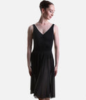 A woman modeling a sleeveless black dance dress with a V-neckline, solid black bodice, and flowing sheer black skirt panels, photographed against a light gray studio background.