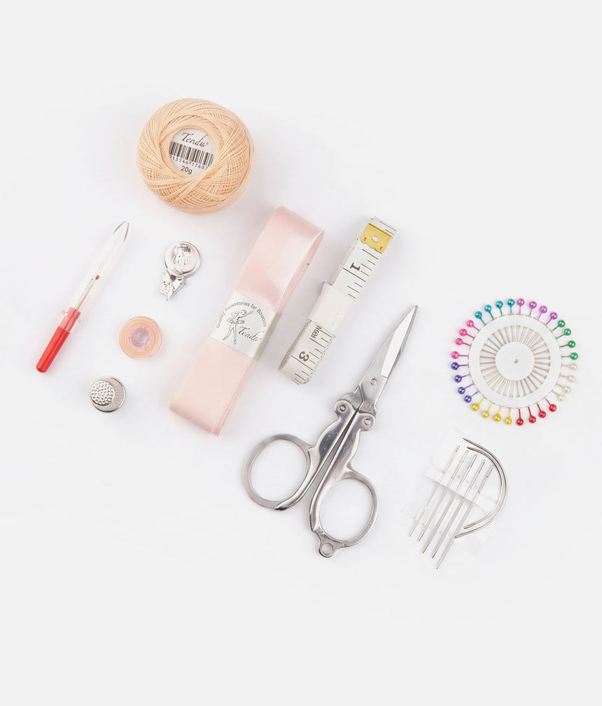 Tendu sewing kit contents arranged on white, including pink ribbon and thread, scissors, tape measure, pins, thimble, seam ripper and straight and curved needles.