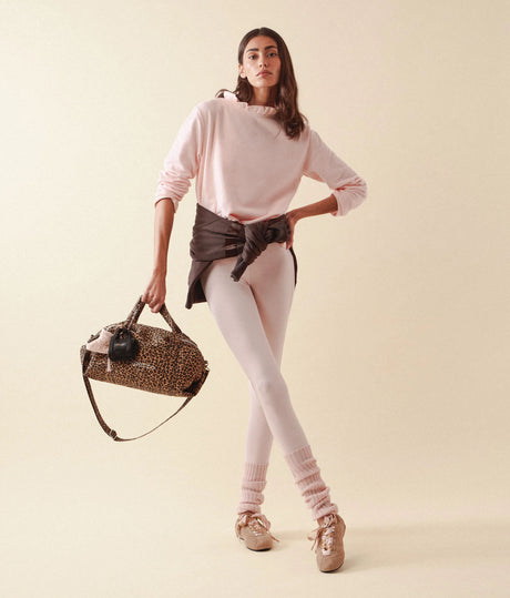 A model stands against a warm beige studio background wearing a light pink ruffled long-sleeve top, pale pink leggings, pink knit leg warmers, and beige sneakers. She is holding a leopard-print duffel bag with a black strap and a black belt tied around her waist.