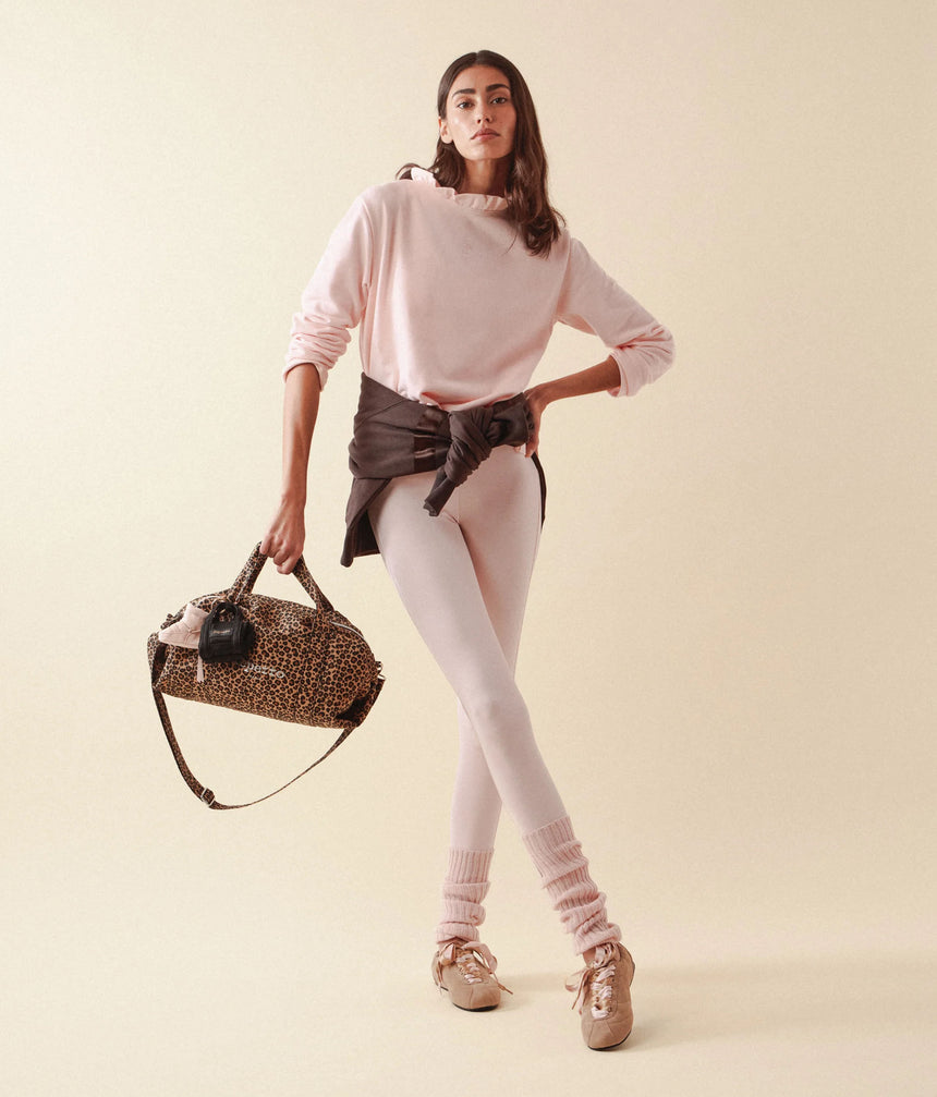 A model stands against a warm beige studio background wearing a light pink ruffled long-sleeve top, pale pink leggings, pink knit leg warmers, and beige sneakers. She is holding a leopard-print duffel bag with a black strap and a black belt tied around her waist.