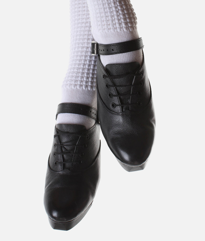 Front view of Antonio Pacelli Essential black leather jig shoes showing laces and straps over white poodle socks