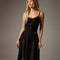 A blonde model wearing a black spaghetti-strap midi dress with a lace bodice, sheer mesh side panels, and a pleated skirt, photographed against a neutral gray studio background.