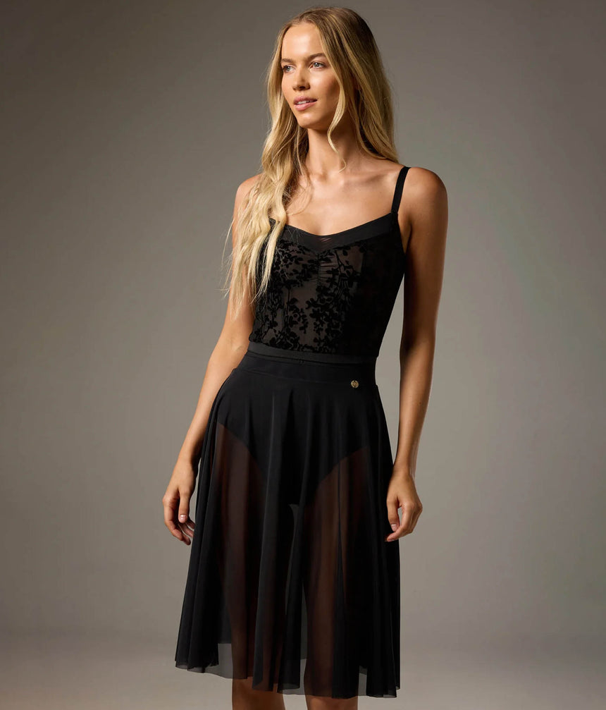 A blonde model wearing a black spaghetti-strap midi dress with a lace bodice, sheer mesh side panels, and a pleated skirt, photographed against a neutral gray studio background.