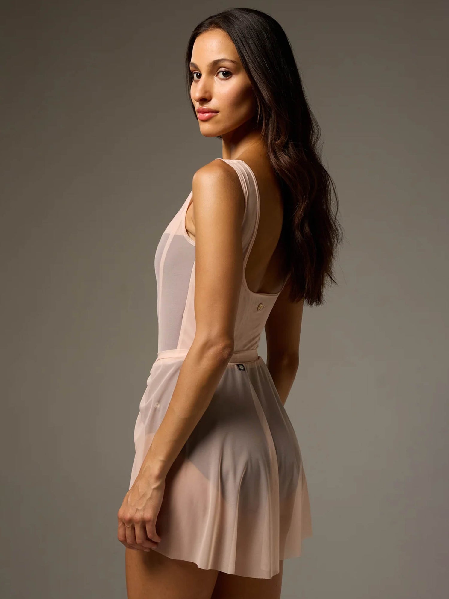 A woman modeling a pale blush pink sleeveless mini dress with a sheer mesh bodice and a flared skirt, photographed against a plain taupe-gray studio background.