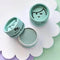 Three small round cosmetic jars of mineral eyeshadow arranged on a white scalloped tray. One jar is open, showing a pale mint green eyeshadow pan, while the other two are closed with light aqua lids featuring a simple black face illustration and the text 'mineral eyeshadow' and 'eyeshadow'.