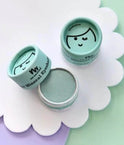 Three small round cosmetic jars of mineral eyeshadow arranged on a white scalloped tray. One jar is open, showing a pale mint green eyeshadow pan, while the other two are closed with light aqua lids featuring a simple black face illustration and the text 'mineral eyeshadow' and 'eyeshadow'.