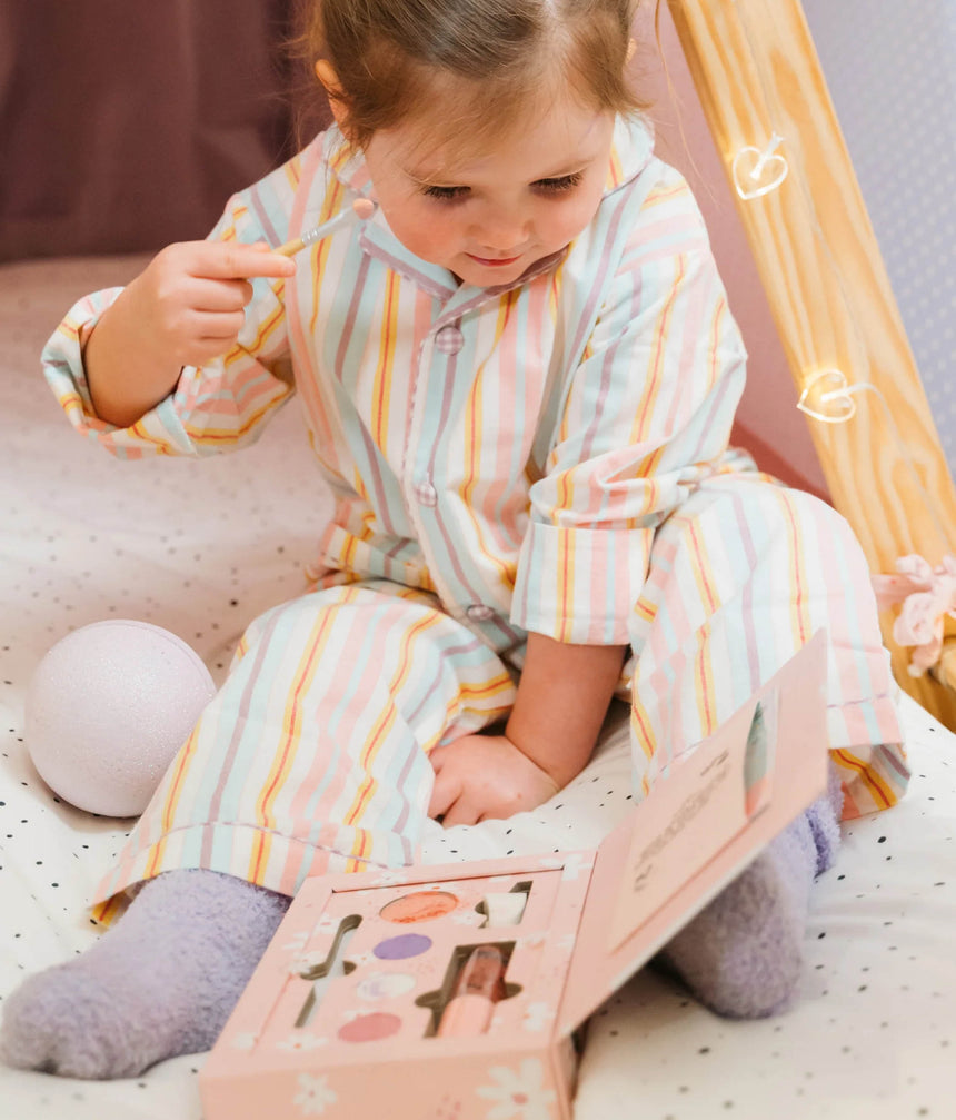 A toddler sits on a speckled cream rug in a play tee, wearing pastel striped pajamas and interacting with a pink makeup palette.