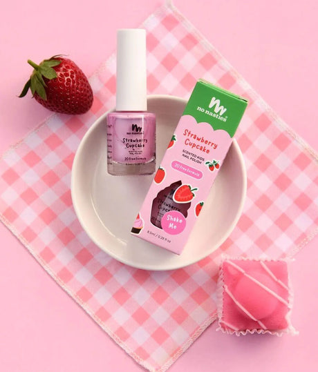 A pink nail polish bottle labeled 'Strawberry Cupcake' next to its green and pink packaging box, styled on a pink-and-white checkered napkin with a strawberry and a pink wrapped candy on a pink background.