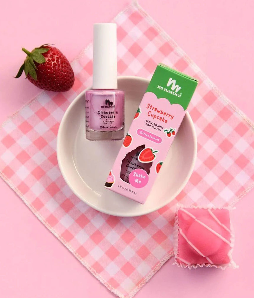 A pink nail polish bottle labeled 'Strawberry Cupcake' next to its green and pink packaging box, styled on a pink-and-white checkered napkin with a strawberry and a pink wrapped candy on a pink background.