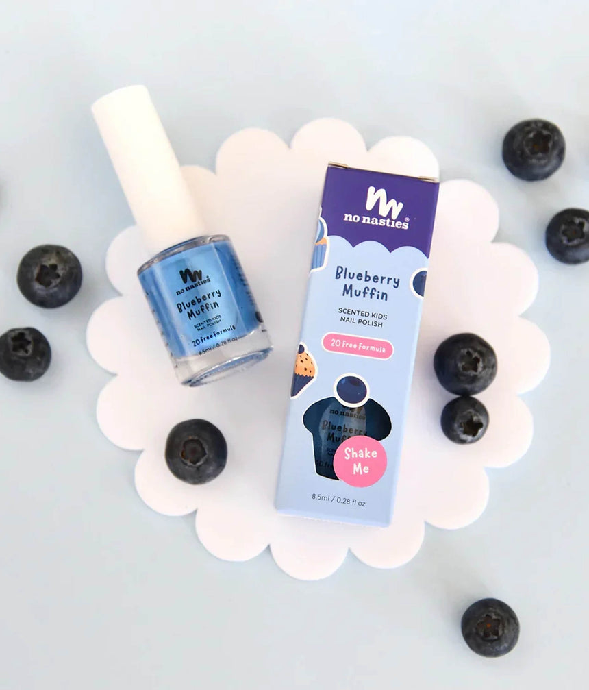 A bottle of No Nasties Blueberry Muffin scented nail polish and its matching retail box, styled on a white scalloped plate with scattered blueberries on a light blue background.