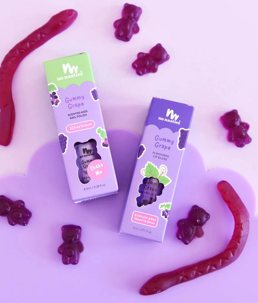 Two boxes of No Marigold Gummy Grape lip gloss and scented hair wax are displayed on a purple heart-shaped plate, surrounded by loose purple and red gummy bears and wavy gummy candies on a light purple background.