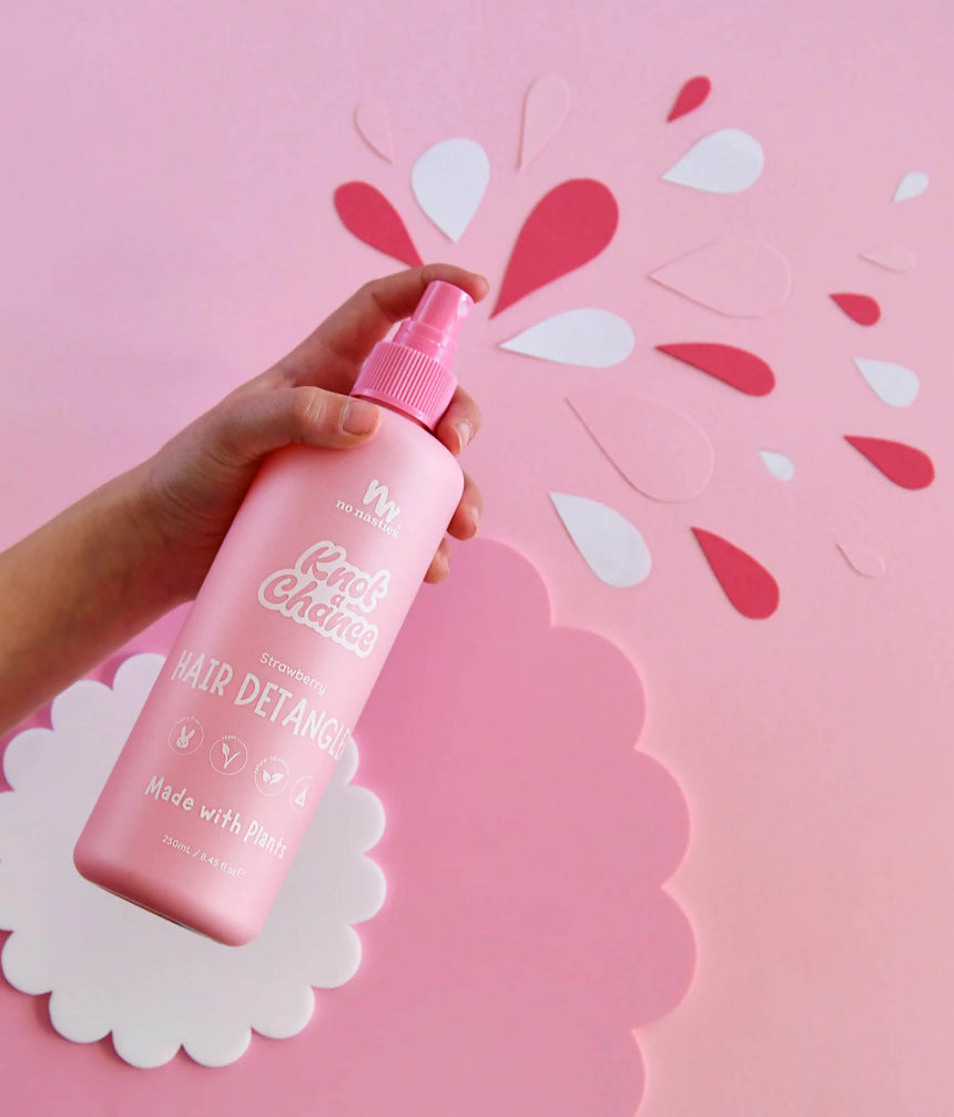 Hand holding No Nasties Strawberry Hair Detangler in a pink spray bottle against a pink paper splash background
