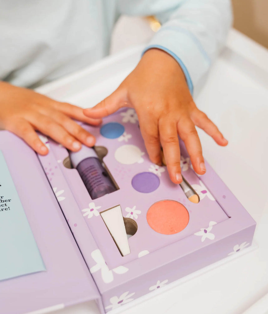 A child's hands resting on a lavender makeup gift set with white floral patterns, containing a purple tube, a blue circular makeup pan, a purple circular makeup pan, and a peach circular makeup pan.
