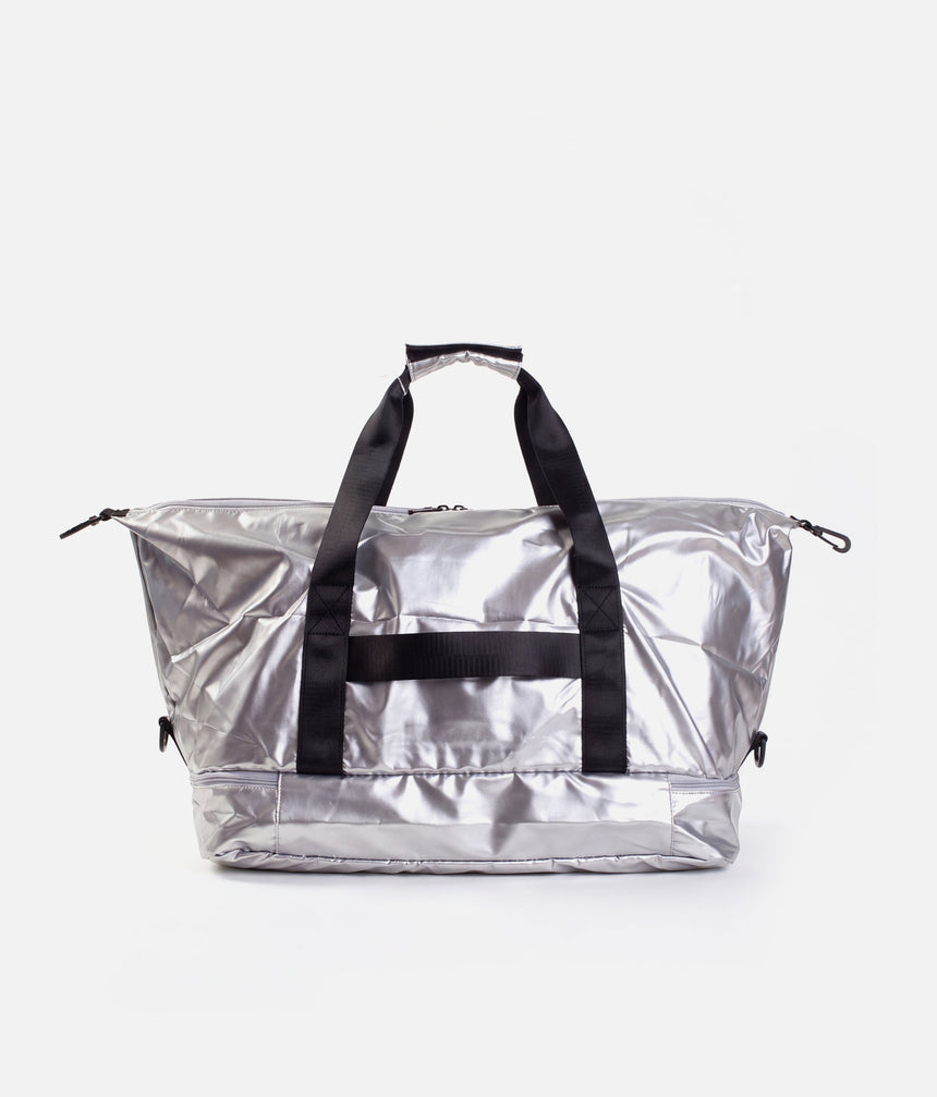 A metallic silver duffel bag with black handles and a black horizontal strap detail, shown against a plain light gray background.
