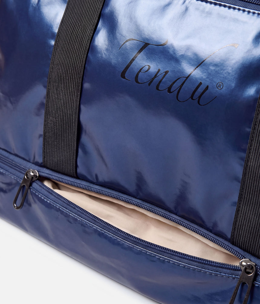 Close-up of a shiny navy blue duffel bag with black webbing handles and a black zipper pocket showing a light beige interior. The brand name 'Tendu' is printed in black script on the front.