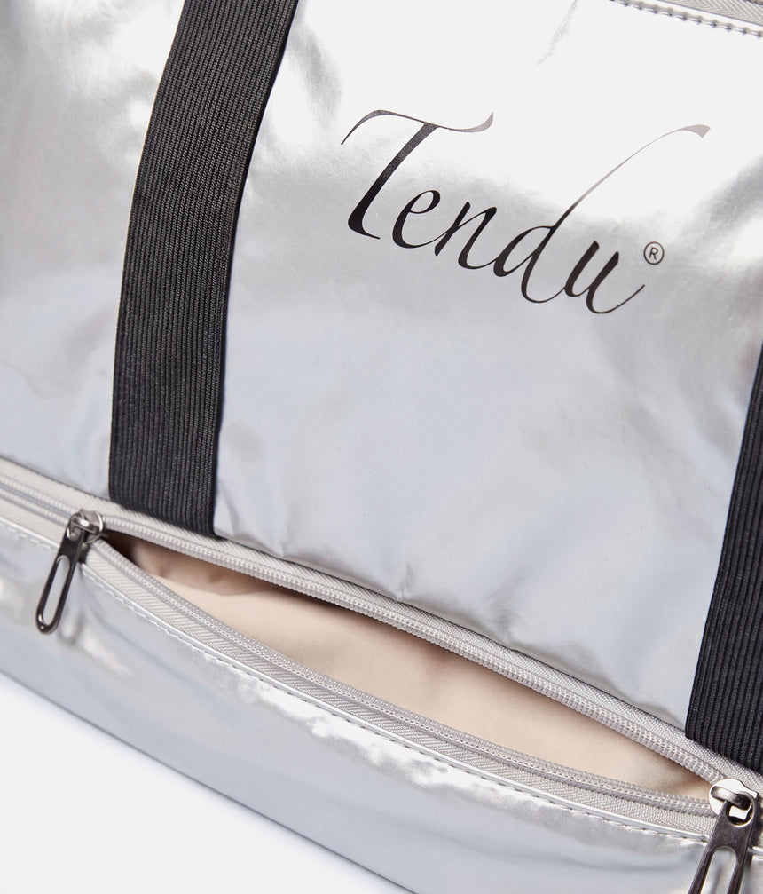 Close-up of a metallic silver duffel bag with black elastic side straps and a partially unzipped front pocket showing a beige lining. The brand name 'Tendu' is printed in black script on the front.